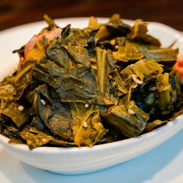 A white bowl filled with dark green collard greens and bits of red ham on a white plate.